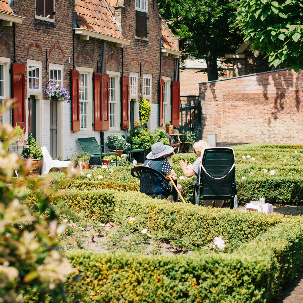 Zwei ältere Damen sitzen auf Gartenstühlen zwischen den niedrigen Hecken des Vorgartens eines ehemaligen Bauernhauses