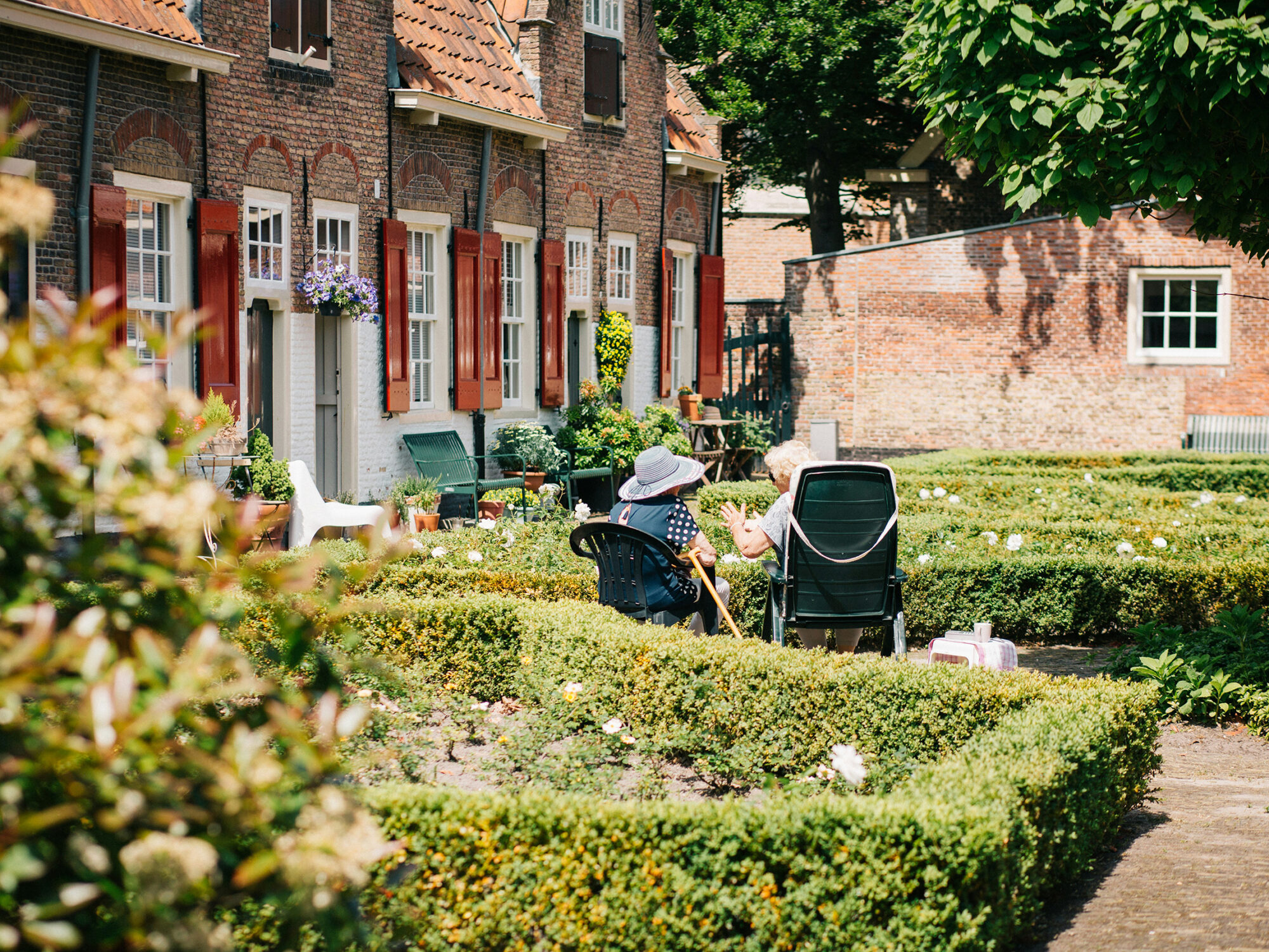 Zwei ältere Damen sitzen auf Gartenstühlen zwischen den niedrigen Hecken des Vorgartens eines ehemaligen Bauernhauses